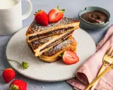 Gestapelte French Toast mit Nutella, bestäubt mit Puderzucker und garniert mit frischen Erdbeeren. Ein Glas Milch und eine Schüssel Nutella im Hintergrund.