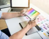 Projektant wybiera kolory z palety na tablecie, korzystając z niego jak ze słownika poligraficznego. Obok leży laptop i wydrukowane wzorniki.