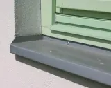 Szary parapet zewnętrzny zamontowany przy oknie z zielonymi okiennicami. Idealny moment, kiedy montować parapety zewnętrzne, to po ociepleniu elewacji.