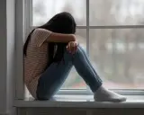 Dziewczyna siedzi skulona przy oknie, ukrywając twarz w ramionach. Jej postawa sugeruje głęboki smutek, być może po traumatycznym przeżyciu, jak gwałt.