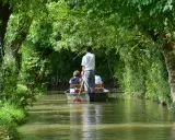 Une petite barque navigue sur un canal bordé d'arbres luxuriants. Un homme debout manœuvre la barque avec une rame, transportant des passagers.