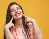 Une jeune femme au sourire éclatant, doigts pointés vers ses joues, semble expliquer la signification d'un sourire pincé.
