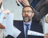 Zaskoczony mężczyzna w okularach, otoczony przez współpracowników z dokumentami, czuje się jakby miał **dwa etaty**.