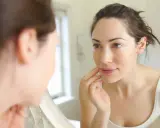 Une femme examine son visage dans un miroir, touchant sa joue. Sa peau semble très fine, peut-être signe d'une carence.