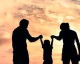 Siluetas de una familia jugando bajo un cielo anaranjado. Un niño es levantado por sus padres, un ejemplo de aprendizaje social y emocional.