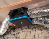 Unité de ventilation maison avec conduits argentés, installée sous un plafond en bois et au-dessus d'un mur en pierre.