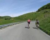 Ciclisti in salita su una strada panoramica, con verdi colline e cielo azzurro. Un'avventura che sembra un **passo cento croci**.