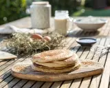 Tortitas de avena recién hechas, apiladas en una tabla de madera. Ingredientes como huevos y leche sugieren un desayuno casero.