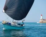 Un voilier bleu avec une grande voile noire navigue près d'un phare. Le skipper, portant un t-shirt bleu, semble observer un ovni 25 dans le ciel clair.