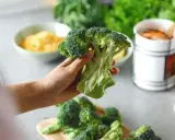 Mano sosteniendo un brócoli fresco, listo para preparar deliciosas recetas de brócoli. En el fondo, queso rallado y zanahorias.