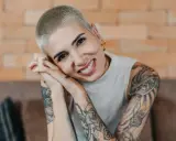 Lächelnde Frau mit Buzz Cut und vielen Tattoos, die ihre Hände vor dem Gesicht verschränkt.