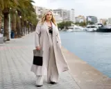 Kobieta o figurze jabłka w eleganckich stylizacjach, spacerująca nad wodą.