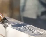 Ręka maluje pędzlem drewnianą ramę okna białą farbą do okien drewnianych na zewnątrz.