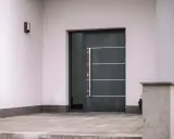 Nowoczesne drzwi wejściowe, ciemnoszare, z długą, metalową klamką i bocznym przeszkleniem. Czy to drzwi stalowe czy aluminiowe?