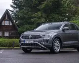 Szary Volkswagen T-Roc używany, zaparkowany przed drewnianym domkiem w lesie. Idealny na wypady za miasto.