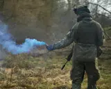 Żołnierz w kamuflażu odpala niebieski granat dymny, przestrzegając zasad ASG.