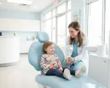 Uśmiechnięte dziecko u dentysty, trzymające pluszową zabawkę, siedzi na fotelu, podczas gdy dentystka pokazuje mu lusterko.