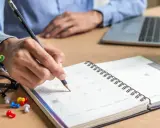 Ręka pisze ołówkiem w kalendarzu, planując dni. Czy to sprawiedliwy grafik w pracy, czy może kolejny dzień pełen obowiązków?