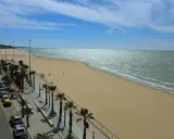 Sonniger Strand mit Palmen und Autos in Sanlúcar de Barrameda. Das Meer glitzert, am Horizont sind Kitesurfer zu sehen.