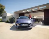 Mujer sonriente junto a un coche alto y morado, aparcado frente a una casa moderna con puertas abiertas.