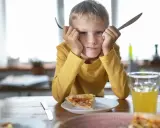 Chłopiec z widelcem i nożem w dłoniach, z miną mówiącą "dziecko nie chce jeść". Przed nim kawałek pizzy i szklanka soku.