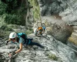 Osoby w kaskach i uprzężach pokonują skalną ścianę, używając liny i karabinków. Cały niezbędny via ferrata sprzęt zapewnia im bezpieczeństwo podczas wspinaczki.