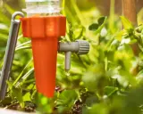 Un système d'arrosage goutte à goutte pour potager, avec une goutte d'eau tombant d'un dispositif orange et gris au milieu de plantes vertes.