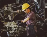 Mujer con casco amarillo y gafas de seguridad trabajando en un motor. Usa guantes negros y un delantal de mezclilla, demostrando el uso de los equipos de protección individual.