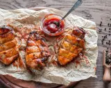 Saftiges Grillfleisch zart machen: Drei gegrillte Hähnchenbrustfilets mit Sauce auf Backpapier, daneben eine Schale Ketchup und eine Holzschaufel mit Pfefferkörnern.