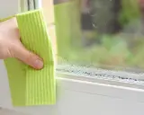 Dłoń z zieloną ściereczką wyciera zaparowane okno, gdzie widać krople wody tworzące linię, przypominającą punkt rosy.
