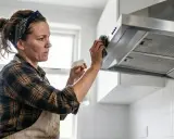 Kobieta w chustce na głowie i fartuchu czyści okap kuchenny. Pokazuje, jak wyczyścić okap z tłuszczu, używając sprayu i gąbki.