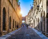 Sonnenbeschienene Gasse in Rhodos Stadt mit Kopfsteinpflaster und historischen Gebäuden. Touristen schlendern durch die mittelalterliche Stadt.