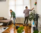 Pareja colaborando en como limpiar la casa: ella aspira el suelo y él quita el polvo de las paredes.