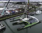 Le trimaran Sodebo Ultim 3, prêt pour la course, est amarré dans un port, entouré de spectateurs et de grues.