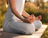 En person sitter i lotusställning på en matta utomhus, med händerna i meditation. Solen lyser genom gräset och skapar en varm atmosfär för morgonmeditation.