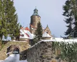 Zamek Czocha zimową porą, otoczony śniegiem i iglastymi drzewami. Kamienny mostek prowadzi do bramy.