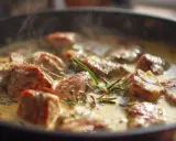 Délicieux sauté de porc mijotant dans une sauce crémeuse, parfumé au romarin. La vapeur s'échappe doucement de la poêle.