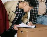 Joven firma contrato de arrendamiento de vivienda, rodeado de cajas.