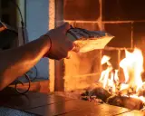 En hand lägger ved i en sprakande brasa. Det syns tecken på soteld i eldstaden.