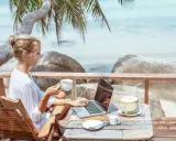Kvinna med kaffe och laptop på en strandterrass, drömmen om ett digitalt nomad jobb.