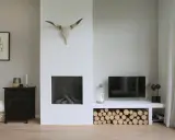 Un salon moderne avec une cheminée blanche épurée, un crâne de taureau, du bois de chauffage et une télévision. Un avant-après réussi pour relooker une cheminée.