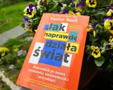 Książka Vaclava Smila "Jak naprawdę działa świat" wśród kwiatów. To przewodnik po przeszłości, teraźniejszości i przyszłości, który pomaga zrozumieć, co to odwyk od błędnych przekonań.
