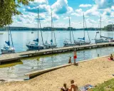 Słoneczny dzień nad jeziorami mazurskimi. Drewniane molo z żaglówkami, piaszczysta plaża z wypoczywającymi ludźmi i błękitne niebo z chmurami.