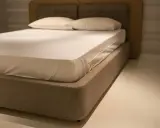 Un lit moderne avec un sommier tapissier, une base recouverte de tissu beige, prêt à accueillir un matelas blanc et des oreillers.