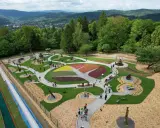 Widok z góry na park z kolorowymi rabatami kwiatowymi i rzeźbami. Widać ścieżki, ławki i ludzi spacerujących po terenie. W tle góry i las.