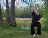 Kvinna i svart klädsel utför yoga på en stol vid en sjö. Naturen är grönskande med träd och vass.