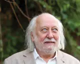 Ritratto di Laszlo Krasznahorkai, scrittore ungherese, con capelli bianchi e barba, sguardo pensieroso rivolto verso l'alto.