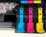 Zbliżenie na mechanizm napełniania tuszy do drukarek. Widoczne są kolorowe klipsy blokujące wkłady: cyjan, magenta i żółty.