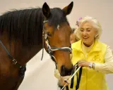 Linda Tellington mit einem braunen Pferd, sie trägt eine gelbe Weste und eine Blume im Haar.