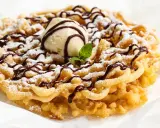 Pyszny funnel cake posypany cukrem pudrem, polany czekoladą i udekorowany gałką lodów waniliowych.